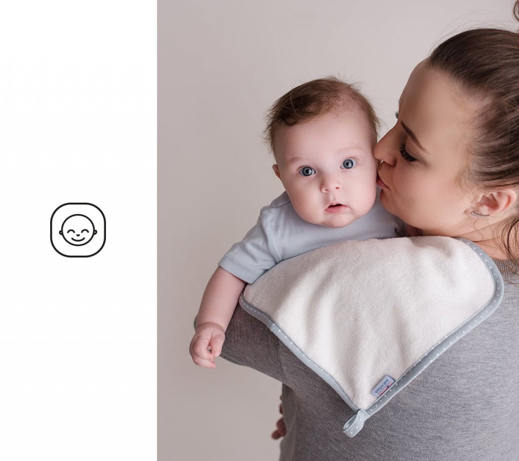 Mama z profilu z długimi, czarnymi rzęsami całuje w policzek dziecko. Dziecko patrzy w obiektyw, ma niebieskie oczy. Na ramieniu mamy spoczywa jasny ręczniczek 