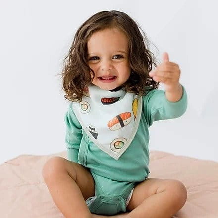 Dziewczynka z ciemnymi włosami siedzi na podłodze, uśmiecha się, a na szyi ma założony śliniak we wzór z sushi