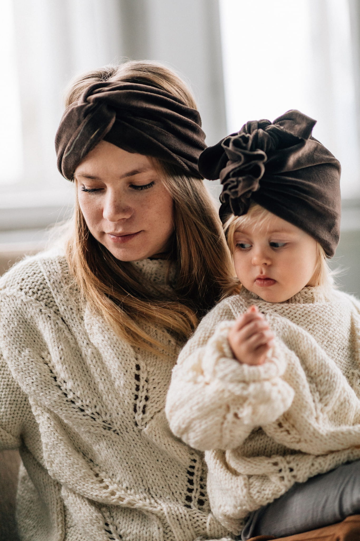 Mama i córka w jasnych swetrach siedzą obok siebie. Mama ma na głowie opaskę, dziecko turban w kolorze ciemnej czekolady