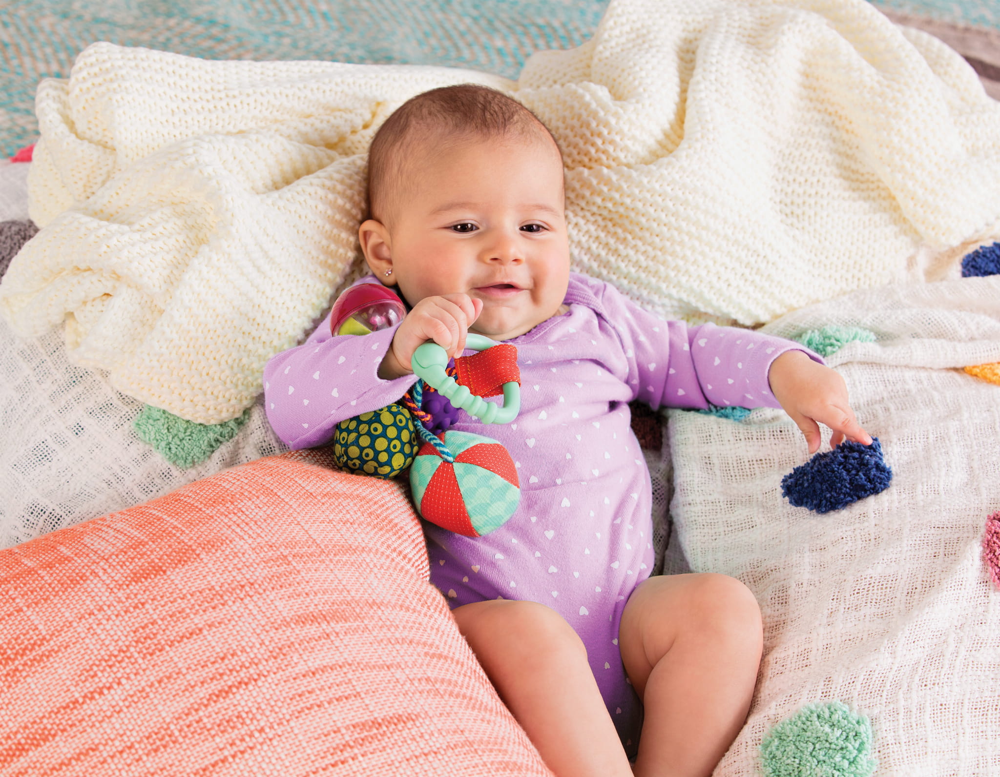 Dziecko w fioletowym body leży na kolorowej pościeli. W uchu ma kolczyk, a w ręku trzyma gryzaka zawieszkę