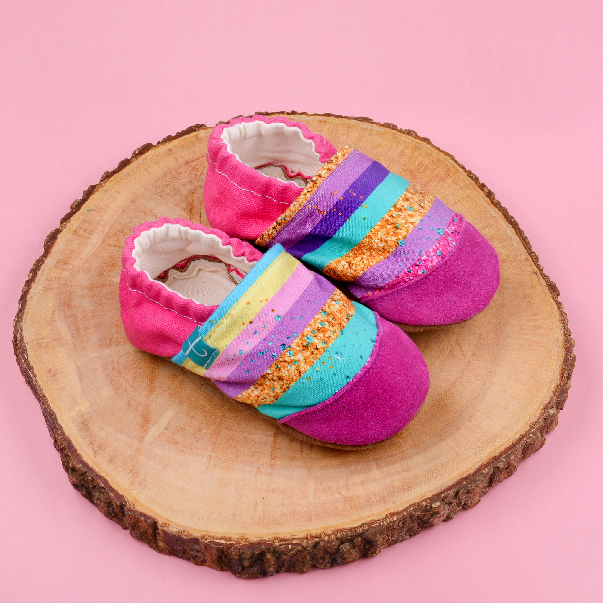 Buty-kapcie w kolorach tęczy i z brokatem postawione na kawałku pnia 