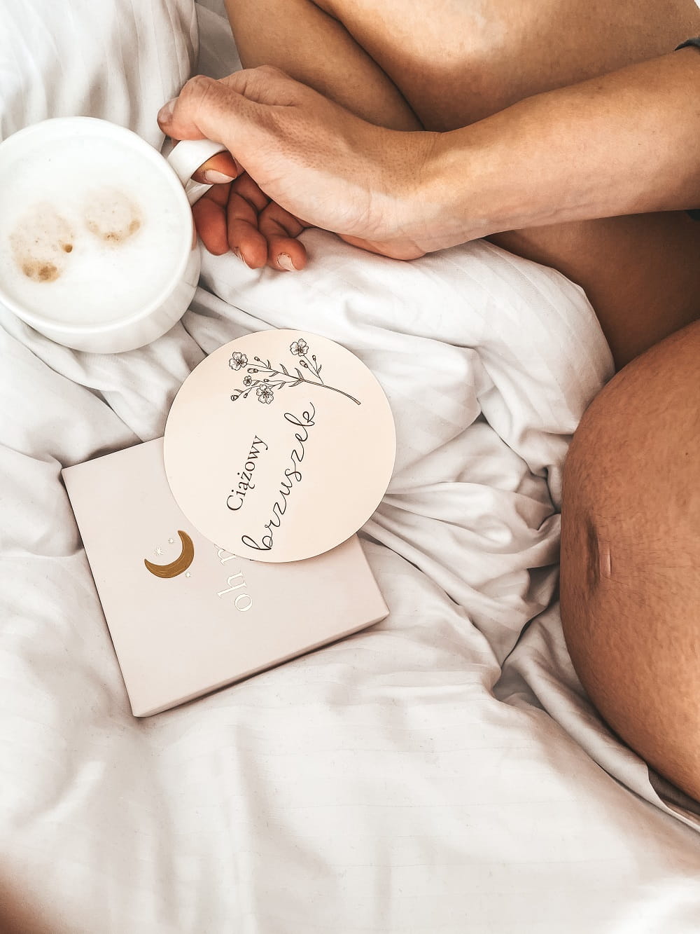 Mama w ciąży siedzi na łóżku i trzyma dłonią kubek z kawą. Przed nią leżą karty do zdjęć