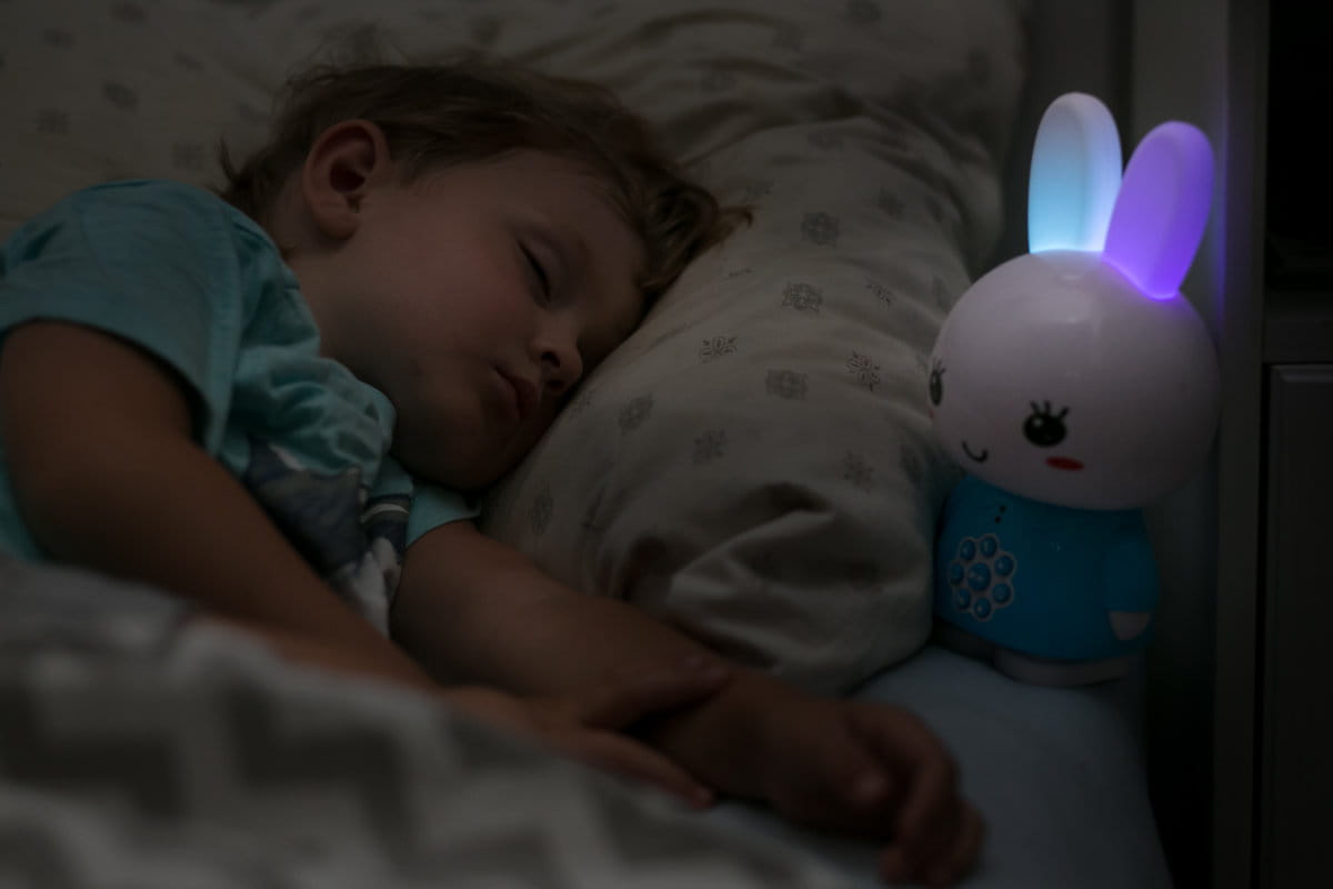Dziecko śpi w łóżku a obok niego, na stoliku nocnym stoi królik ze świecącymi uszami