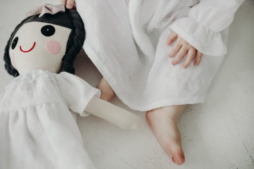 Lalka z czarnymi włosami w białej sukience leży obok stópki dziewczynki w białej sukience
