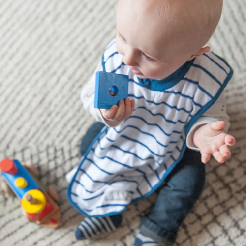 Dziecko z jasnymi włosami siedzi na podłodze. Ma założony długi śliniak w niebieskie paski, a w rączce trzyma niebieski, drewniany klocek