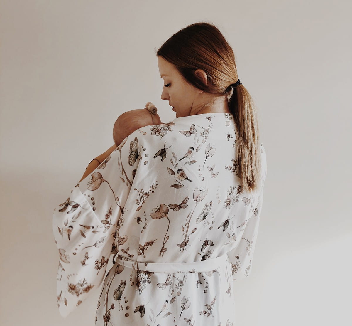Mama z dzieckiem w ramionach stoi tyłem, ubrana jest w szlafrok w kwiatowy wzór