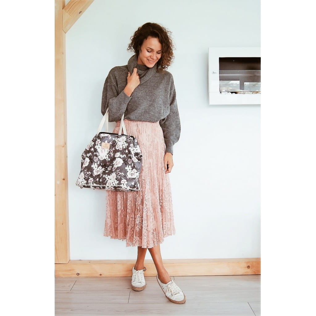 Kobieta w długiej, różowej spódnicy i szarej bluzce trzyma na przedramieniu torbę w kwiatowy wzór
