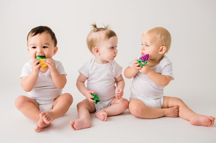 Trójka dzieci w białych bodziakach - dwóch chłopców i dziewczynka - siedzi na podłodze. W rączkach trzymają gryzaki owoce