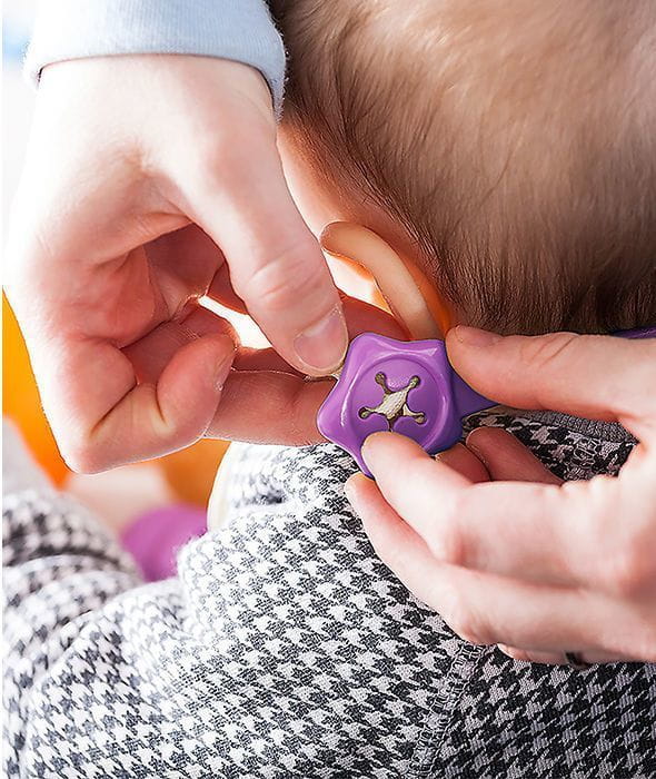 Dłonie zapinają fioletowy uchwyt na śliniaku. Na zdjęciu widać też fragment ucha dziecka