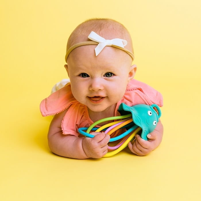 Na żółtym tle leży dziewczynka w opasce z kokardką. W rączkach trzyma grzechotko-gryzak meduzę. Uśmiecha się