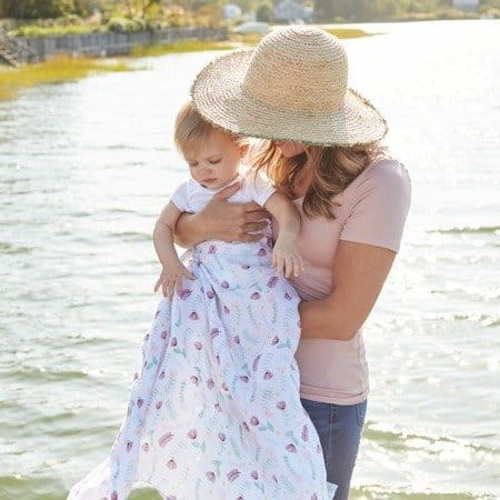 Mama w kapeluszu w słoneczny dzień trzyma otulone otulaczem dziecko. W tle widać wodę