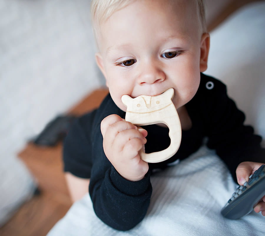 Dziecko w ciemnej bluzeczce z długim rękawem leży na łóżku i gryzie drewniany gryzak