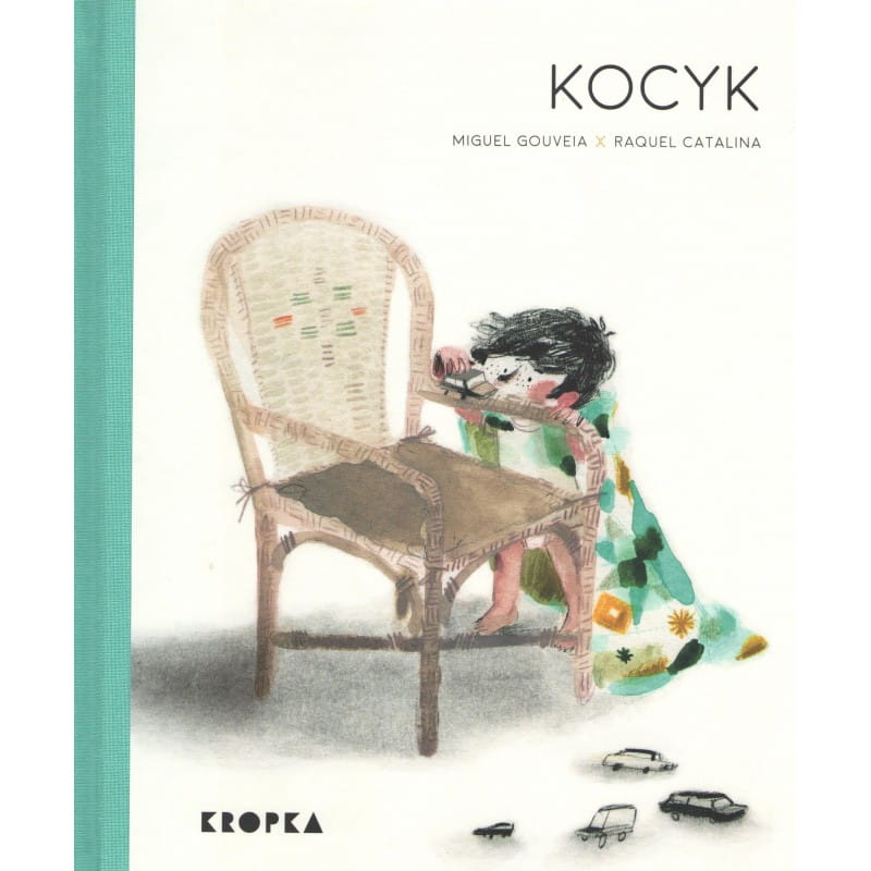 Okładka książki pt. Kocyk - chłopiec w kocyku założonym na plecy niczym peleryna spogląda na bujany fotel