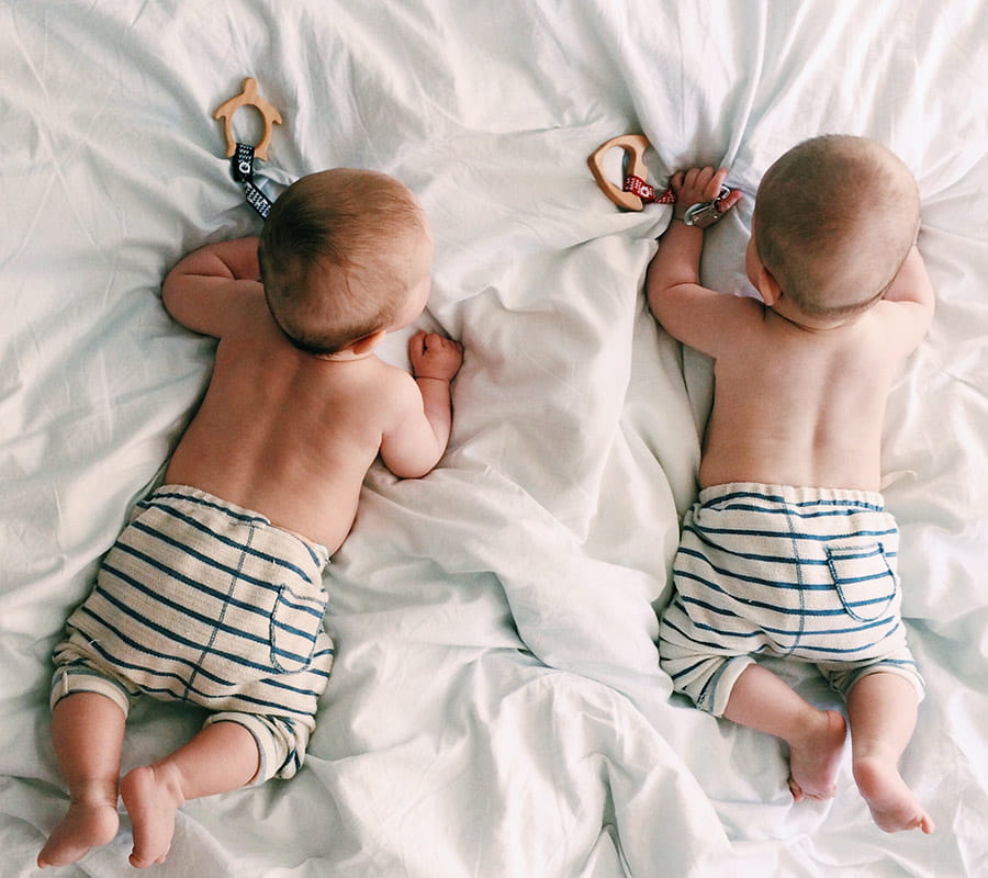 Dwoje dzieci w spodenkach w paski i z gołymi pleckami leży na łóżku na brzuchach. W rączkach trzymają zawieszki, do których przyczepione są drewniane gryzaki - żółwik i rybka